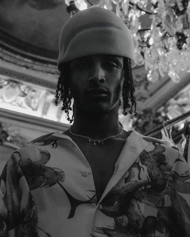 Model in a printed jacket and pale bucket hat beneath a chandelier