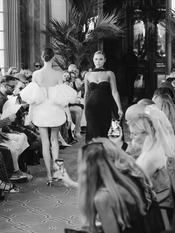 The runway seen from behind: a white feathered dress passing seated guests