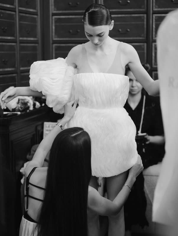 A dresser adjusting a white feathered mini dress backstage